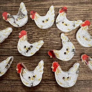 May include: A collection of white chicken-shaped buttons with red combs and wattles, featuring black and yellow speckled details. Each button has two holes for sewing, with a glossy finish and a distinct curved silhouette resembling a hen.