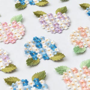 May include: A collection of embroidered hydrangea flower patches in various pastel colors including pink, blue, purple, and peach. Each patch features clusters of small, multi-petaled flowers with white centers and green stitched leaves.
