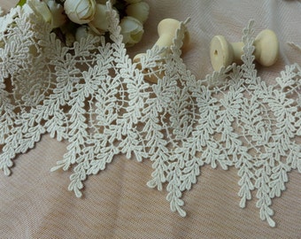 May include: An off-white lace trim featuring a repeating botanical leaf pattern with intricate scalloped edges. The lace is displayed alongside two small, light-colored wooden spools and a cluster of small, pale artificial flowers.