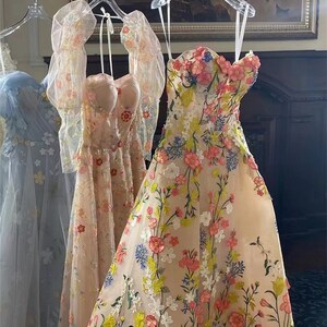 May include: Two sheer, floral embroidered dresses on hangers. The dress on the left is light blue with a floral pattern. The dress on the right is beige with a floral pattern in shades of pink, yellow, and green.
