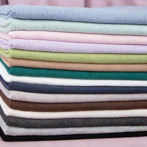 May include: A collection of folded ribbed knit fabric in a range of colours. Shades include pale blue, mint green, pink, lilac, light green, beige, teal, navy, white, brown, grey, and black. The fabric displays a textured finish.