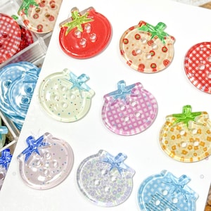 Puede incluir: Una colección de botones redondos con forma de tomate que presentan cuatro agujeros en el centro. Los botones muestran varios patrones, incluyendo cuadros vichy rojos y blancos, pequeños estampados florales y colores sólidos, con un tallo verde o azul.
