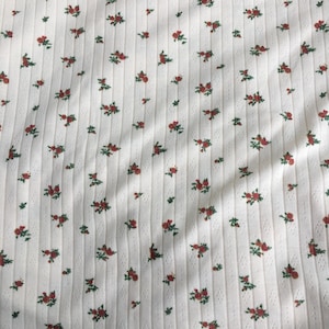 May include: White ribbed fabric with a repeating pattern of small red roses and green leaves. The fabric has a textured, vertical striped design, creating a subtle visual interest. The floral pattern is evenly distributed across the surface.