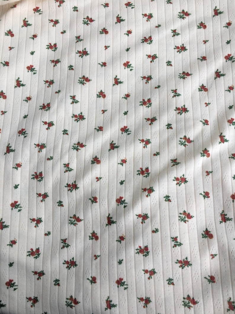 May include: White ribbed fabric with a repeating pattern of small red roses and green leaves. The fabric has a textured, vertical striped design, creating a subtle visual interest. The floral pattern is evenly distributed across the surface.