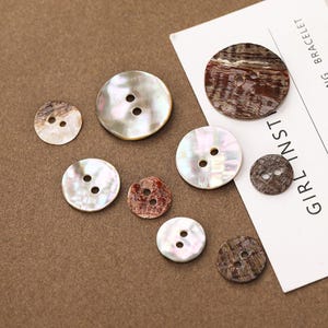 May include: A collection of round, two-hole buttons in varying sizes. The buttons feature iridescent white, pearlescent, and dark brown wood-grain patterns. A white card with the partial text "GIRL INST" and "G BRACELET" is visible.