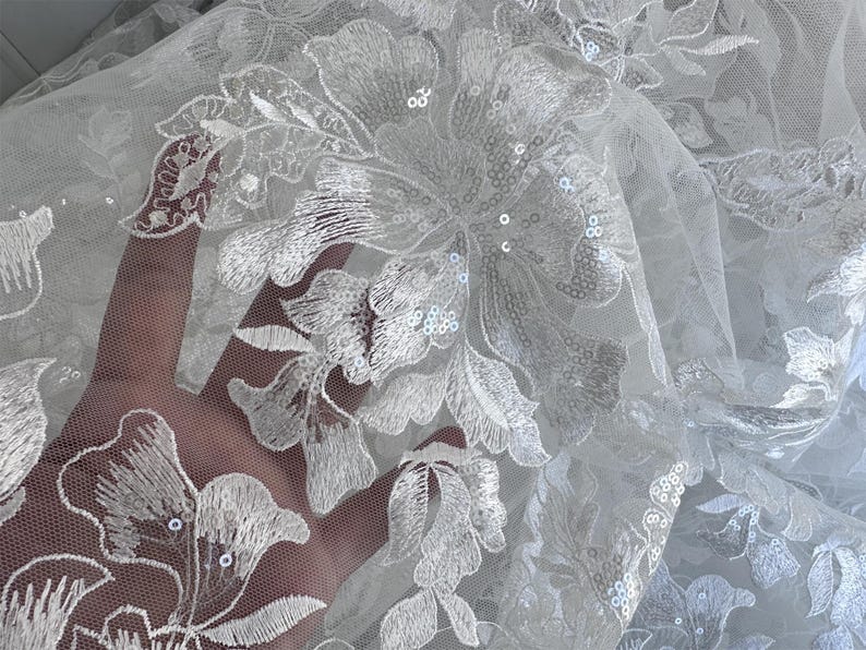 May include: White floral lace fabric with sequins. The lace features a delicate floral pattern with a sheer mesh background.