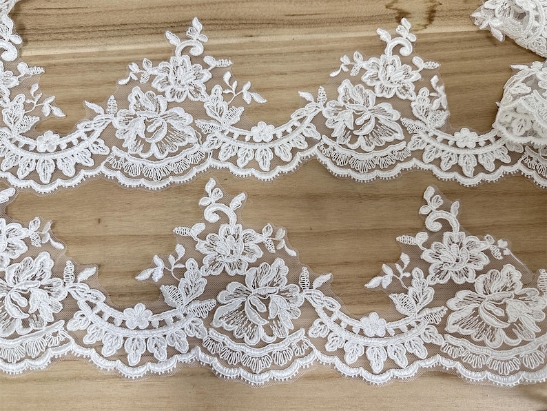 May include: Close-up of white floral lace trim. The lace features a repeating pattern of flowers, leaves, and scalloped edges. The delicate design is set against a sheer background, ideal for bridal or craft projects.