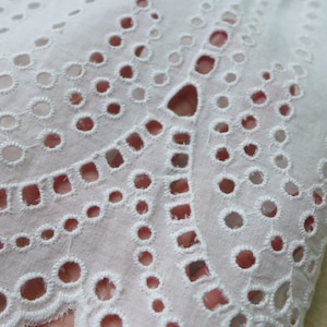 May include: White cotton fabric with a delicate eyelet pattern. The fabric has a scalloped edge and is ideal for crafting projects.