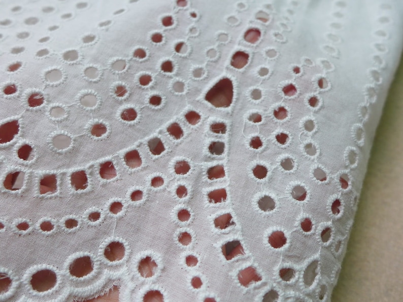 May include: White cotton fabric with a delicate eyelet pattern. The fabric has a scalloped edge and is ideal for crafting projects.