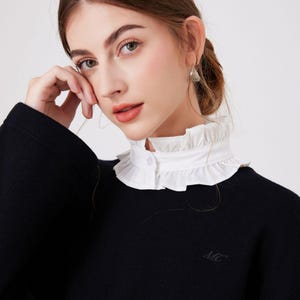 May include: A black sweater with a white, ruffled collar. The collar is buttoned at the front. The sweater has the initials "MC" embroidered on the lower left side.