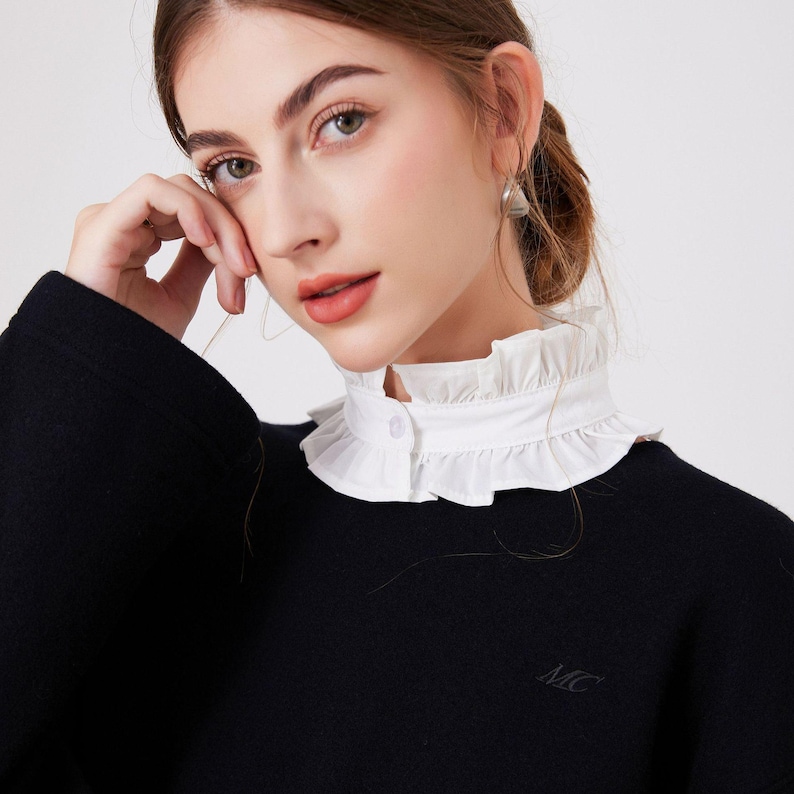 May include: A black sweater with a white, ruffled collar. The collar is buttoned at the front. The sweater has the initials "MC" embroidered on the lower left side.