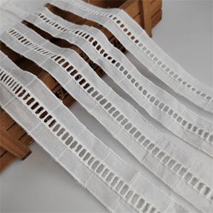 May include: Close-up of several white eyelet lace ribbons. Each ribbon features a series of rectangular cut-out designs. The ribbons are arranged diagonally, showcasing their texture and pattern. Ideal for sewing, crafting, and adding a decorative touch to various projects.