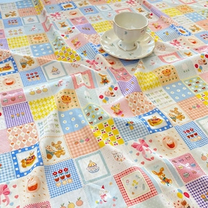 Könnte beinhalten: Ein Patchwork-Tischdecke mit verschiedenen pastellfarbenen Quadraten. Jedes Quadrat zeigt ein anderes Design, darunter Teddybären, Kirschen, Cupcakes und Schleifen. Eine weiße Tasse und Untertasse liegen auf der Tischdecke.