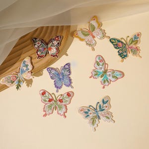 May include: A collection of embroidered butterfly patches in various colors. The patches feature detailed floral designs in shades of pink, green, blue, and purple. The butterflies are arranged on a light beige surface.
