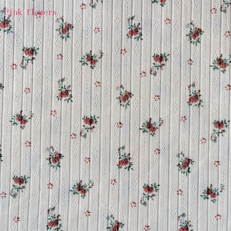 May include: Cream-colored fabric with a ribbed texture and a repeating floral pattern. The pattern features clusters of red roses with green leaves and small red dots. The words "Pink flowers" are in the top left corner.