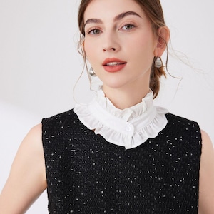May include: A sleeveless black top with a white ruffled collar. The collar is buttoned at the front. The top has a textured appearance with small, reflective embellishments. The model is wearing silver earrings.