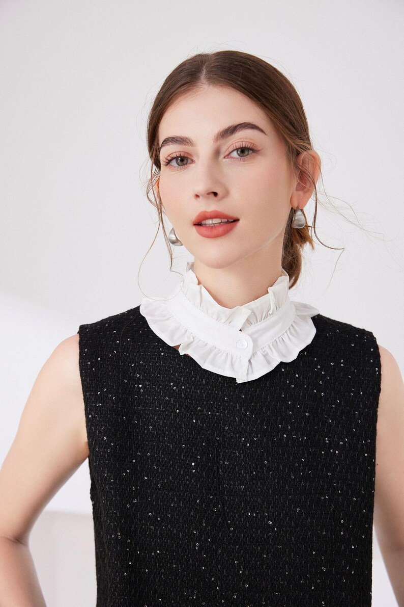 May include: A sleeveless black top with a white ruffled collar. The collar is buttoned at the front. The top has a textured appearance with small, reflective embellishments. The model is wearing silver earrings.