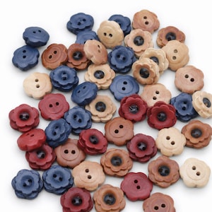 Puede incluir: Una colección de pequeños botones con forma de flor en varios colores, incluyendo azul, rojo, marrón y tonos de madera natural. Cada botón presenta un borde festoneado y dos agujeros centrales para coser, esparcidos sobre un fondo blanco.