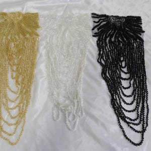 Puede incluir: Tres ribetes de flecos de cuentas en dorado, blanco y negro. Cada ribete presenta un diseño de cuentas en la parte superior y hebras de cuentas en cascada.