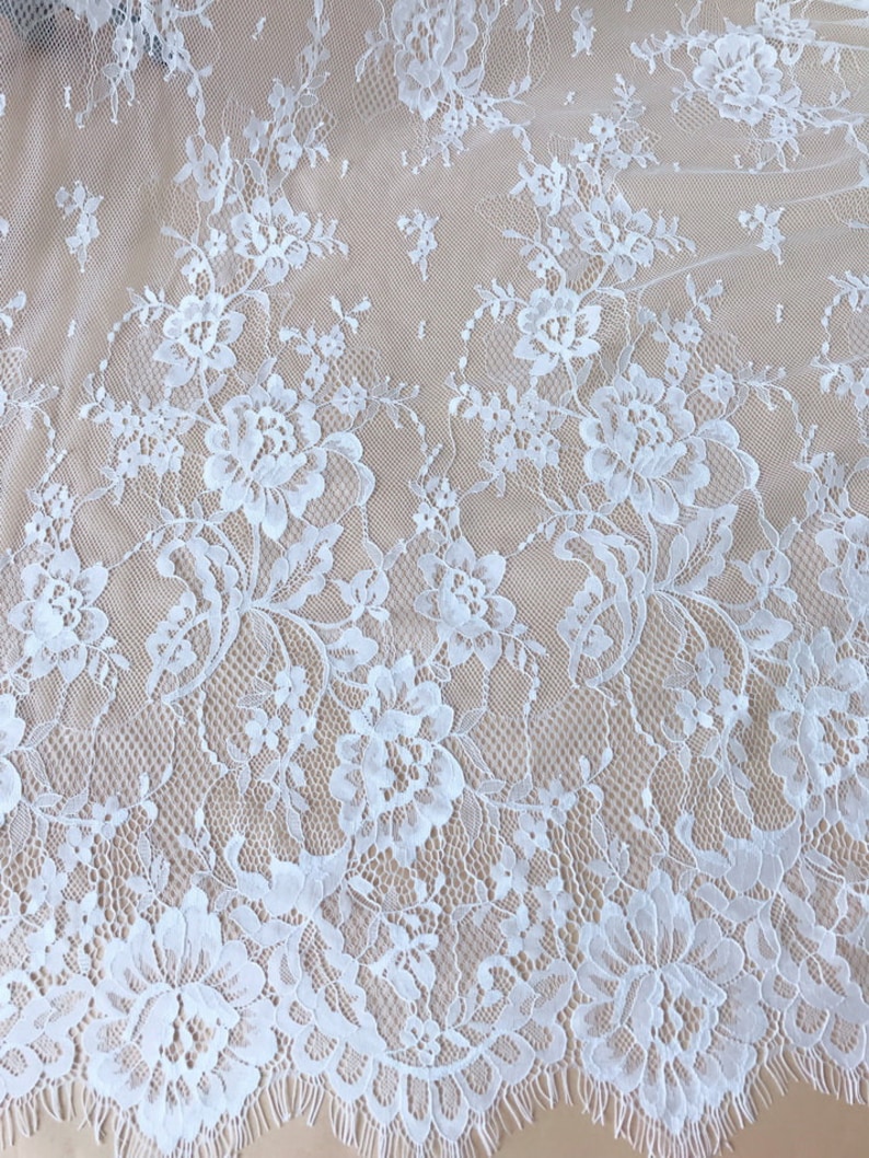 May include: White lace fabric with a delicate floral pattern. The lace has a scalloped edge and a soft, flowing texture.