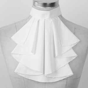 May include: A white ruffled jabot collar, shown on a mannequin. The collar has a high, structured neckline and multiple layers of cascading ruffles, giving a voluminous and elegant look. The fabric seems to be a crisp, lightweight material.