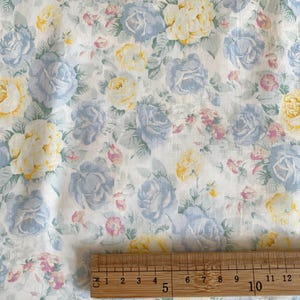 May include: Floral fabric featuring a pattern of blue, yellow, and pink roses on a white background. The fabric is measured with a wooden ruler, showing a length of approximately 10 inches.
