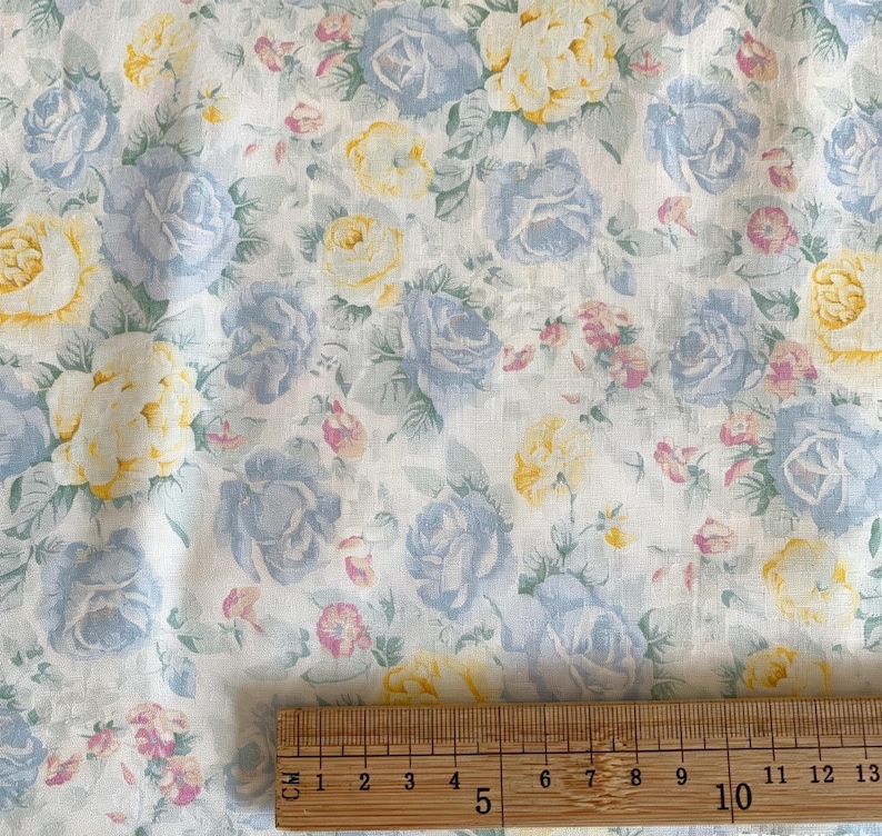 May include: Floral fabric featuring a pattern of blue, yellow, and pink roses on a white background. The fabric is measured with a wooden ruler, showing a length of approximately 10 inches.