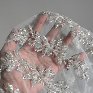 May include: Close-up of delicate white lace fabric decorated with silver beads and sequins. The intricate floral design is visible against the sheer background. The lace is suitable for bridal wear or special occasion garments.