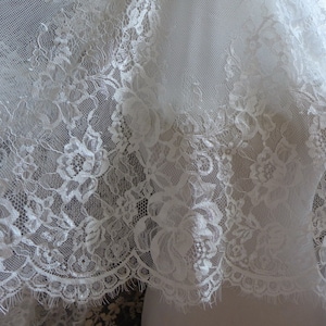 May include: White lace fabric with a delicate floral pattern. The lace has a scalloped edge and is soft and sheer.