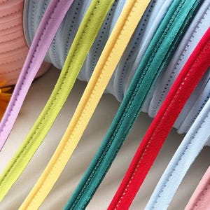 May include: A collection of multi-coloured piping trim strips featuring a soft, suede-like texture and a central stitched line. The strips are arranged in a parallel row, showcasing shades of lavender, lime green, yellow, dark teal, red, and light blue.