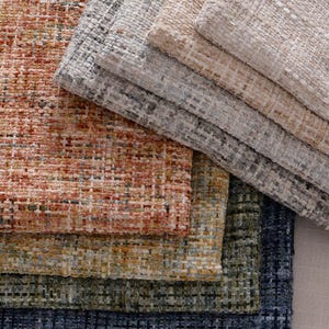 May include: A selection of textured fabric samples in a range of colours. The colours include shades of orange, grey, beige, green, and blue. The fabric has a woven appearance and a slightly rough texture.