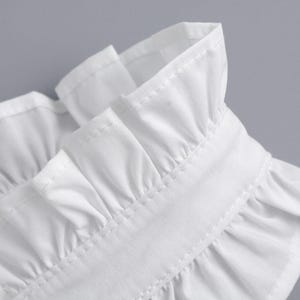 May include: Close-up of a white garment collar with ruffled edges. The collar is made of a crisp, white fabric and features detailed stitching. The design suggests a formal or vintage style.