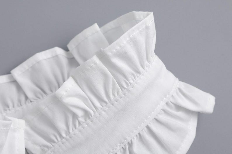May include: Close-up of a white garment collar with ruffled edges. The collar is made of a crisp, white fabric and features detailed stitching. The design suggests a formal or vintage style.