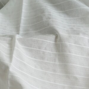 May include: White cotton fabric with a subtle striped pattern. The fabric is wrinkled and has a soft, textured feel.