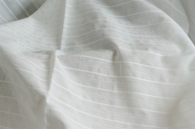 May include: White cotton fabric with a subtle striped pattern. The fabric is wrinkled and has a soft, textured feel.