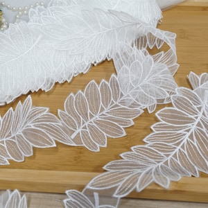 May include: White lace trim featuring a delicate leaf design. The trim is displayed on a wooden surface.