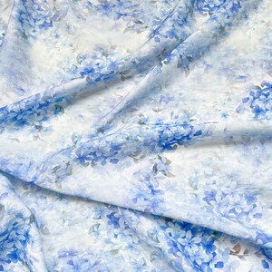 May include: A lightweight fabric featuring a delicate floral pattern with clusters of blue and light blue blossoms scattered across a white background. The material has soft folds and drapes, creating a textured appearance with varied shades of blue.