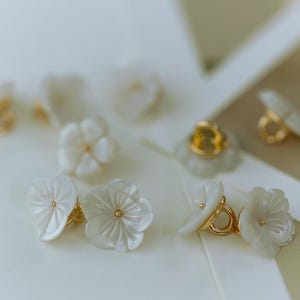 May include: White flower-shaped earrings with gold-coloured accents. The earrings feature a delicate, layered petal design and a small gold centre. The earrings are set against a soft, neutral background.