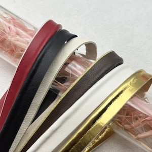 Könnte beinhalten: Eine Sammlung flacher Lederstreifen in Rot, Schwarz, Weiß, Dunkelbraun und Metallic-Gold, die in einem geschichteten Stapel angeordnet sind. Die Streifen haben eine glatte, strukturierte Oberfläche und liegen neben einem transparenten Röhrchen mit rosa Trockengras.