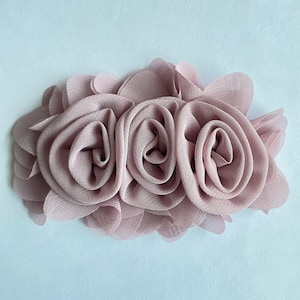 May include: A cluster of three blush pink fabric roses, crafted from a soft, layered material. The roses are arranged in a horizontal row, creating a delicate and feminine design.