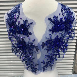 May include: A pair of royal blue lace appliques featuring intricate floral embroidery, embellished with matching sequins, beads, and round pearls. The decorative pieces are crafted on a sheer blue mesh fabric, designed for garment ornamentation.