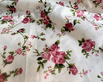 May include: White fabric with a repeating pattern of pink roses and green leaves. The roses vary in size and are clustered together in some areas. The fabric has a slightly wrinkled texture.