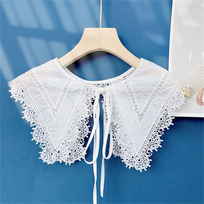 Puede incluir: Un cuello de encaje blanco, desmontable, con un delicado dise&ntilde;o floral y cierre de lazo. El cuello se muestra en una percha de madera sobre un fondo azul. El intrincado dise&ntilde;o del cuello a&ntilde;ade un toque de elegancia.