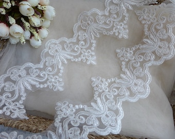 Puede incluir: Primer plano de un ribete de encaje blanco con borde festoneado. El encaje presenta un diseño floral y de hojas, con delicados detalles bordados. El ribete se coloca sobre una tela beige claro, con un ramo de flores blancas en el fondo.