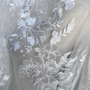 May include: White floral lace fabric with a delicate pattern of flowers and leaves. The lace is embellished with small sequins, adding a touch of sparkle.