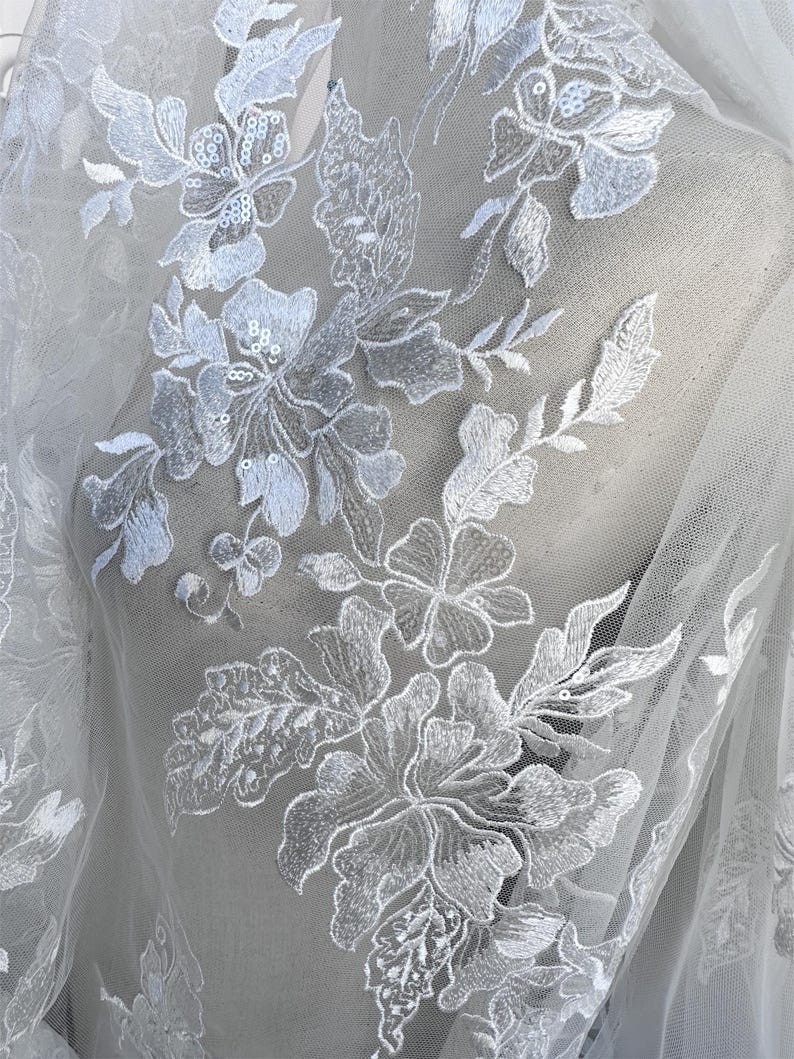 May include: White floral lace fabric with a delicate pattern of flowers and leaves. The lace is embellished with small sequins, adding a touch of sparkle.