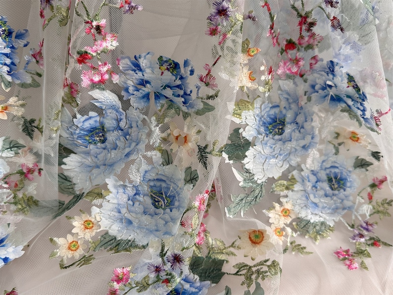 Puede incluir: Tela de tul blanco con patrones florales bordados en azul y verde. Las flores son grandes y detalladas, con un efecto delicado, casi como de acuarela.