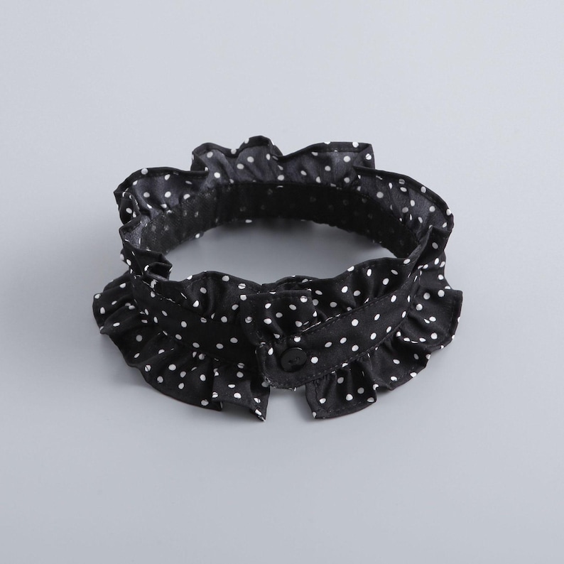 May include: Black and white polka dot ruffled collar with a button closure. The collar is made of a lightweight fabric and features a delicate, ruffled edge. The white polka dots are evenly spaced across the black background.