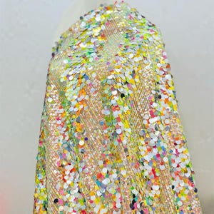 May include: A vibrant fabric sample covered in iridescent sequins. The sequins are in a variety of colors, including silver, yellow, pink, and blue. The fabric is draped over a mannequin, showcasing its texture and drape.