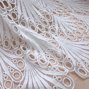 May include: White lace fabric with a detailed peacock feather design. The fabric features a repeating pattern of open circles and curved lines, creating a delicate and airy texture. Ideal for crafting or apparel.